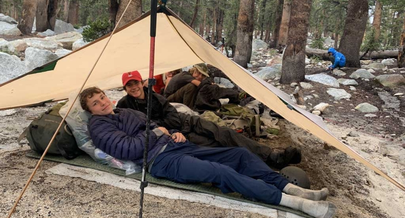 People lie resting under a tarp shelter in a wooded area